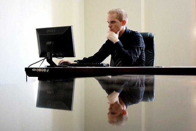 homem de roupa preta utilizando um PC em uma sala. O homem está pensativo, com a mão no queixo